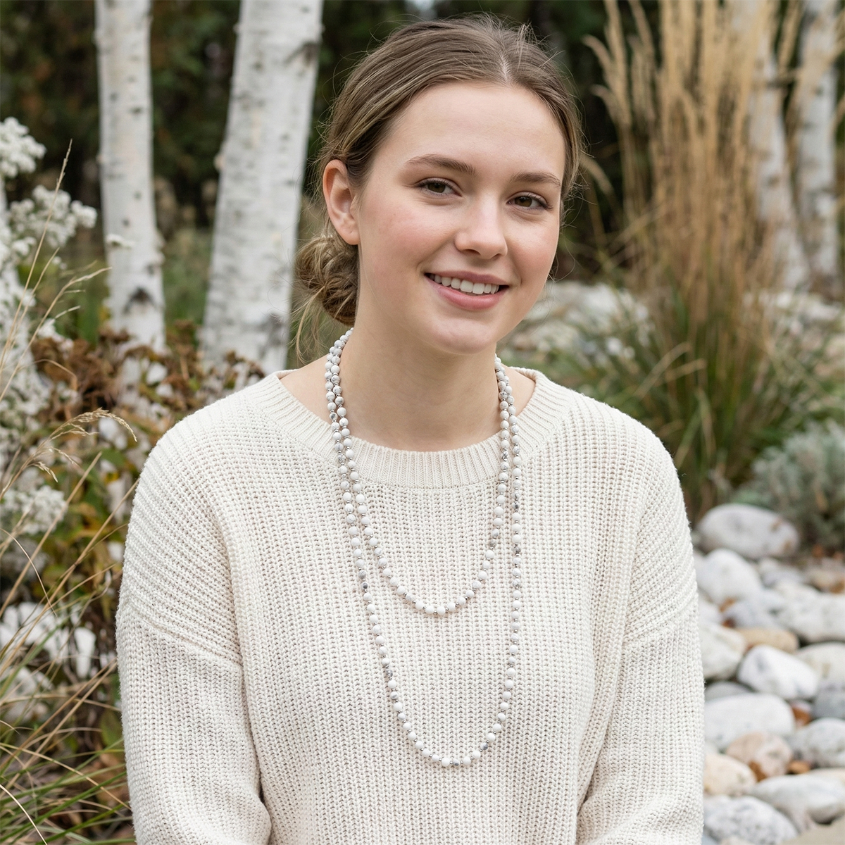 Woman wearing a white sweater and beaded necklace in a natural setting with trees and rocks.