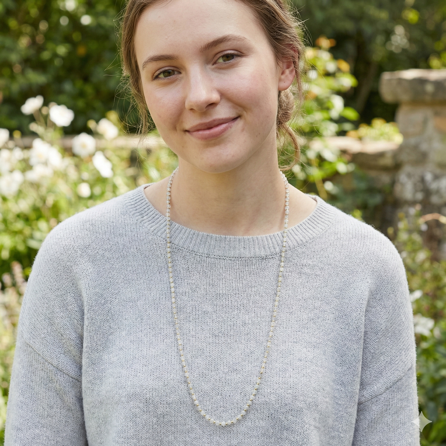 Woman wearing a gray sweater and long necklace outdoors with greenery in the background