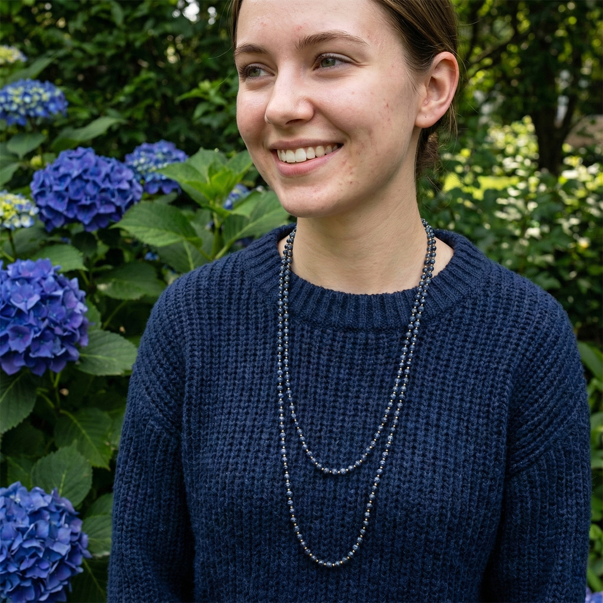 Woman wearing a navy sweater with layered necklaces in front of blue flowers