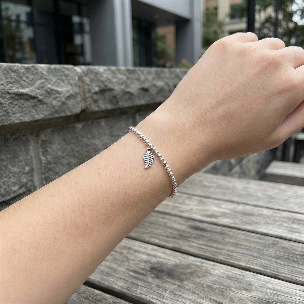 Hand wearing a bracelet with a leaf charm on a wooden surface with a stone wall background