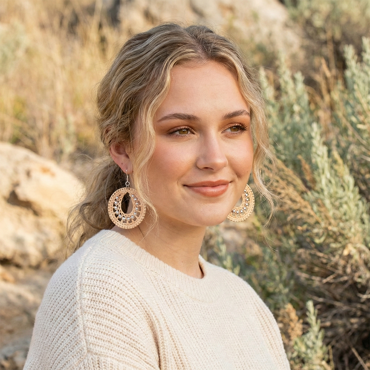 Woman with large hoop earrings standing in a natural setting with rocks and plants.